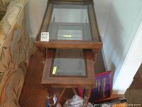Two wooden framed nesting tables with glass tops shown stacked with the smaller table nesting under the larger one.