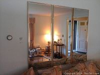 Photo of three rectangular beveled wall mirrors standing upright against a wall with gold trim reflecting living room furniture and light