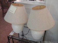 Pair of off-white ceramic lamps with beige fabric shades placed on glass table, showing full side view.