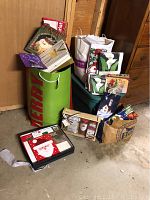 Photo shows large green gift bag labeled 'MERRY' alongside multiple smaller white and decorative holiday gift bags and boxes, some with snowman and Christmas motifs, placed on the floor against a wooden wall.