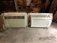 Two window air conditioning units placed side by side on concrete floor in storage with window installation kits attached. One has a simple dial interface, the other with controls on the front panel.