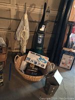 Wide shot of Eureka carpet cleaner and Sears floor polisher standing upright against garage wall with cord wrapped around carpet cleaner, wicker hamper filled with cleaning items and Black & Decker cleaning set box on top, other items seen nearby.