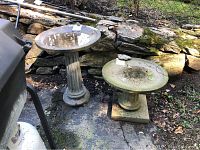 Two cement bird baths outdoors next to stone rock wall; one larger circular basin on fluted column with round base, one smaller circular basin on fluted column with square base.