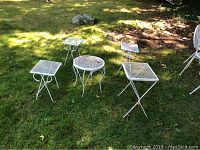 Five white painted wrought iron patio tables arranged on grass outdoors, showing sizes and designs with visible rust and wear.