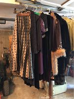 Photo showing garments hanging on rack including the long gray polka dot skirt, black glittery dress, brown embroidered coat with faux fur cuffs and trim, and long black coat