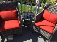 Two gray synthetic wicker chairs with red cushions flanking matching round wicker side table with glass top on deck