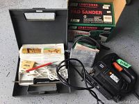 Overview of all items: black carrying case with woodburning tool and accessories, Black & Decker jigsaw, Craftsman pad sander and original box, sanding sheets