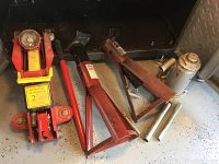 Red hydraulic floor jack with yellow 2 ton capacity label, positioned beside red metal jack stands and metal support bars on garage floor.