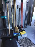 View of shovels, brooms, push brooms, and two boxes of garbage bags on floor against metal cabinet