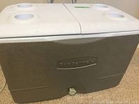 Full side view of the Rubbermaid rolling cooler showing gray body with 'Rubbermaid' embossed on front, white dual lids with cup holders on top, and drainage plug at bottom center.