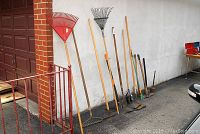 Fourteen assorted garden tools leaning against a wall outdoors, including rakes, shovels, hoes, axes and mallets.