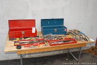 Overall view of both red and blue metal toolboxes on a wooden table, with various hand tools displayed around them including pliers, wrenches, knives, and saw blades.