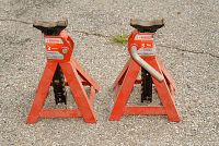 Pair of red metal MotoMaster axle stands positioned upright on pavement with visible labels indicating 3 ton capacity.