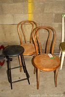 Photo of two wooden bentwood chairs with cane woven seats and a metal base stool with black cushioned seat grouped together against a wall.