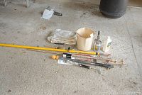 Photo of house painting tools on concrete floor including extension poles, a paint bucket, spray gun, brushes, drop cloths, and bottle.