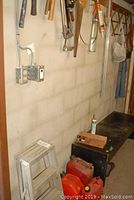 Aluminum step stool beside three red and orange gas cans on the floor, part of a black workbench visible with tools on top and long handles leaning against wall