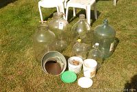 Outdoor photo showing four large glass bottles with two of them having glass stoppers, accompanied by two small containers with corks and some lids, all placed on green grass with white chairs in background.