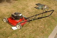 Photo of the lawn mower full side view on dry grass, showing the red deck, black handlebar, and wheels.
