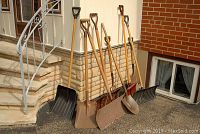 Five shovels leaning against a stone wall, including plastic snow shovels and metal garden shovels with wooden handles, showing wear and rust.