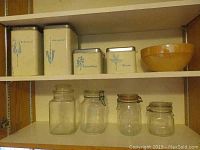 Overview of four vintage cream-colored metal Lincoln Beautyware canisters and orange-brown FireKing mixing bowl on kitchen shelf with four glass jars below.