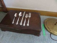 Closed wooden flatware case with a few flatware pieces placed on top, showing the ornate Heritage pattern handles of spoons, forks, and knives.