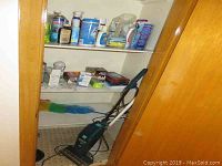 Photo showing blue and black upright Hoover brush vacuum cleaner inside cupboard with shelves holding various cleaning supplies.