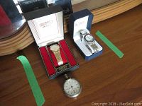 Photo showing the Seiko gold-tone men's quartz wristwatch in its original box, the Activa men's silver-tone wristwatch in its box, and the Elgin silver pocket watch with a white dial and black numerals with a broken minute hand.