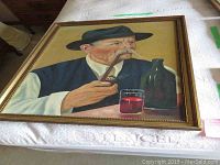 Framed oil painting showing an older man with a blue hat and vest smoking a pipe, with a glass and bottle on table.