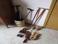 Photo showing two wooden walking sticks with ornately engraved solid brass handles, disassembled parts, five men's large wooden shoe stretchers, adjustable canes, metal trash can, and umbrellas arranged on floor.
