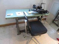 Wide view of large industrial sewing table with black blind stitch sewing machine and rolling chair in front, showing overall setup.