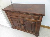 Front view of vintage wood cabinet showing detailed paneling and decorative top edge with one knob missing.