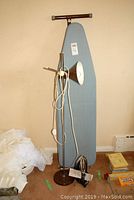 Photo of the blue padded ironing board standing upright, with the retro metal floor lamp in front. The Hamilton Beach steam iron and its manual are on the floor next to the lamp.