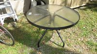 Outdoor round patio table with textured glass top and black metal frame shown from one angle on a grassy outdoor setting.