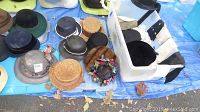 Photo showing a variety of hats laid out on a tarp and inside white plastic containers. Hats include darker colored fedoras, straw hats, and a vintage fur-trimmed hat with floral scarf.
