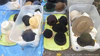 Two large plastic totes filled with assorted vintage hats in various shapes, sizes, and colors including black, beige, green, and white, sitting on blanket or tarp ground covering.