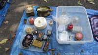 Wide shot showing mixed trinket boxes, porcelain pieces, and crystal glass items arranged inside and outside plastic bins on a blue tarp outdoors