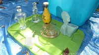 Overall view of assorted vintage glassware and ceramics arranged on a green plastic lid including crystal pitcher, decanters, ceramic bust plant holder, and yellow floral jug
