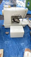 Side view of white Singer sewing machine with foot pedal and detachable cover box on blue tarp.