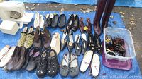 Photo of assorted ladies shoes arranged neatly on a blue tarp, multiple styles including boots, slip-ons, and sneakers, beside a plastic container filled with various sunglasses