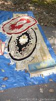View of multiple rugs including a red and beige area rug with floral pattern, a black and beige floral patterned rug, and a rolled blue-green carpet on a blue tarp with scattered leaves around