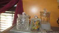 Three clear glass containers with lids, one medium size filled with colorful candies, placed on a surface against an orange wall with a window and purple curtains in background.