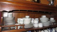 Display of stacks of plates, bowls, and cups on wooden shelf, featuring monogrammed Royal Doulton China with gold trim.