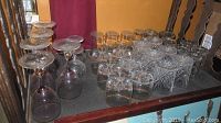 Wide shot of assorted stemware and drinking glasses arranged on a dark surface featuring multiple clear wine glasses and various sized small drinking glasses with cut patterns.