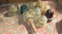 Photo showing ten glass insulators of various colors including clear, aqua blue, amber, and dark brown placed on a patterned rug, one with a wooden handle attached.