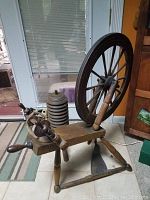 Photo showing antique wooden spinning wheel from side angle with flywheel, treadle, and flyer assembly visible.