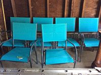 Photo showing six turquoise blue mesh outdoor chairs arranged in two rows. The chairs have dark metal frames and armrests, with mesh fabric seats and backrests.