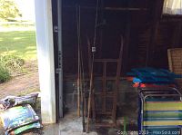 Tapered metal and wood hand truck standing upright against a garage wall with four bathandle fishing poles placed alongside it.