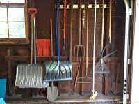 Various long-handled yard tools hanging on a wooden wall in a garage, including shovels, rakes, pitchfork, broom, and an orange plastic scoop shovel.