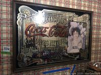 Vintage Coca Cola advertising mirror with visible oxidizing and age patina, showing detailed text and painted portrait of a woman.