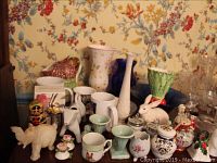 Full display of assorted porcelain and glass items on wood table with floral wallpaper background.